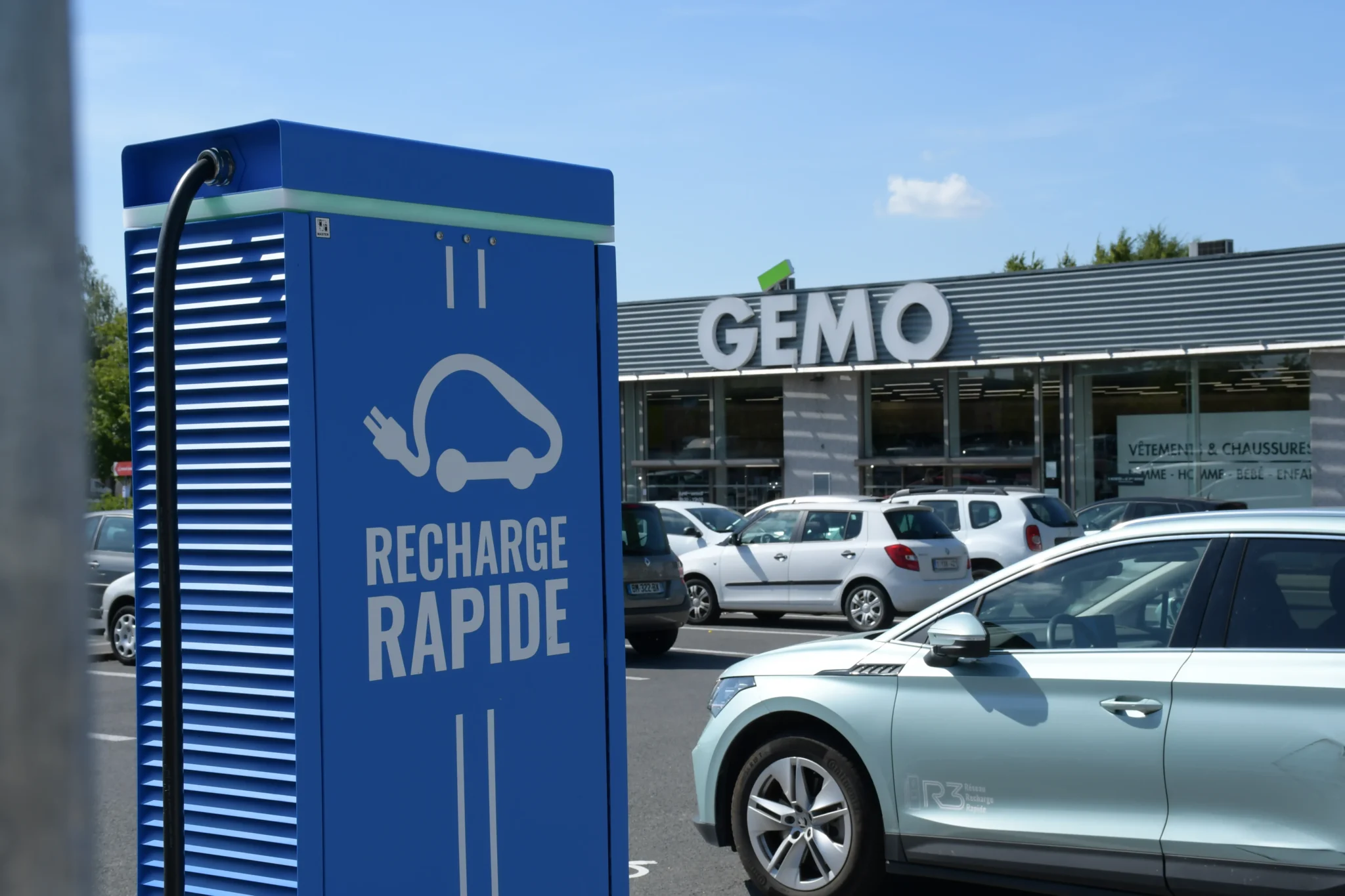 Borne de recharge rapide pour véhicule électrique installée sur le parking d’un centre commercial, voiture électrique stationnée en charge devant un magasin, infrastructure de mobilité durable facilitant la recharge lors des achats