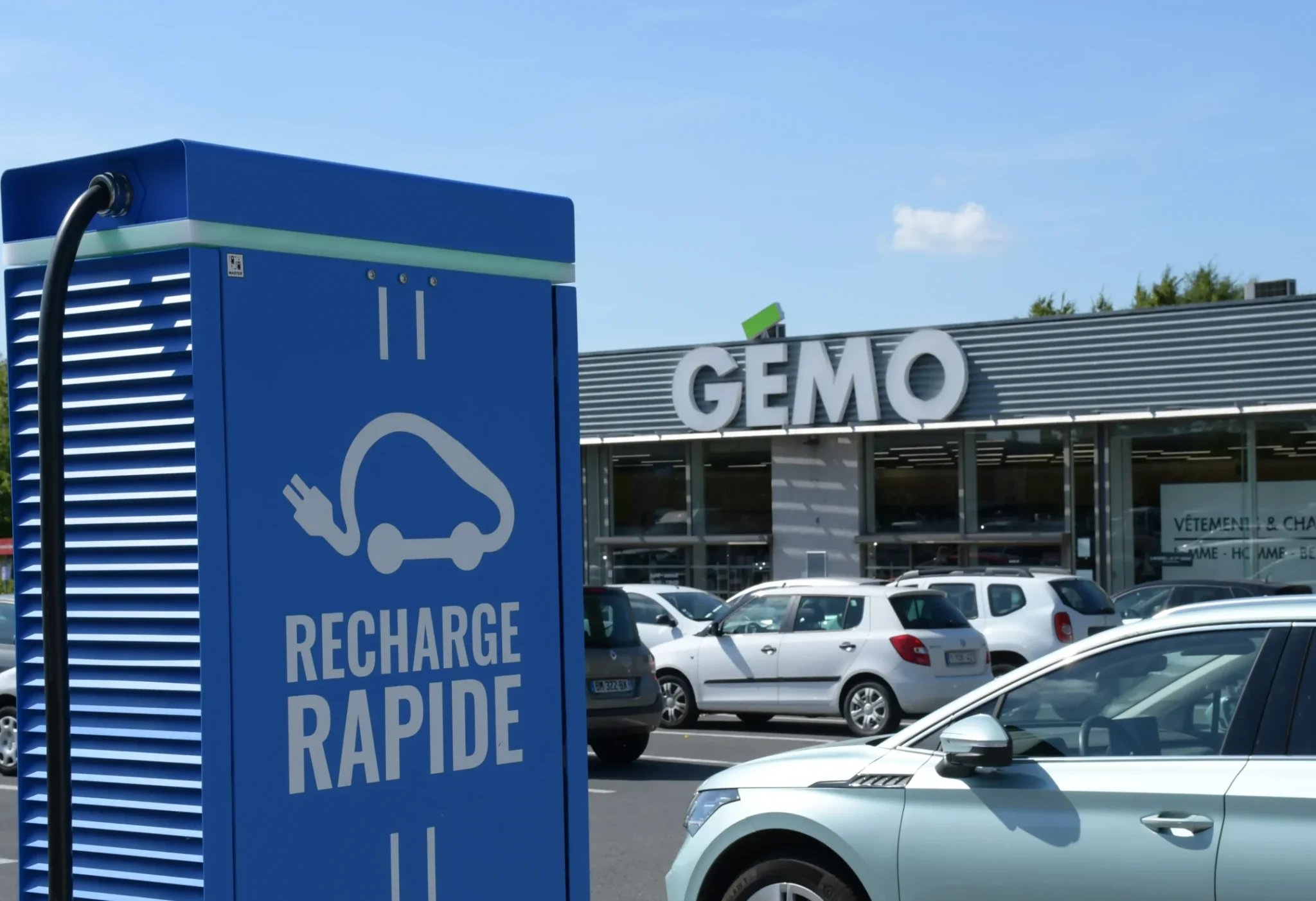 Borne de recharge rapide pour véhicule électrique installée sur le parking d’un centre commercial, voiture électrique stationnée en charge devant un magasin, infrastructure de mobilité durable facilitant la recharge lors des achats