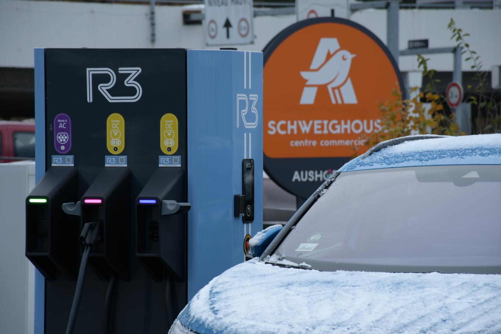 Photo d'une voiture en premier plan avec une borne de recharge R3, par temps d'hiver avec un peu de neige sur le capot de la voiture.