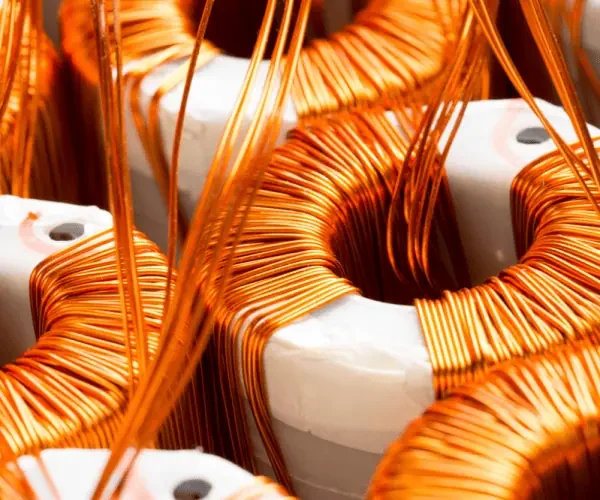 L’image montre un gros plan sur des bobines de fil de cuivre enroulé autour de structures en céramique blanche, probablement utilisées dans des composants électriques tels que des transformateurs ou des inducteurs. Les fils de cuivre brillants, soigneusement enroulés, reflètent une lumière vive, mettant en valeur leur aspect conducteur et leur précision d’assemblage. Cette scène illustre des matériaux essentiels pour la fabrication de dispositifs électriques et électromagnétiques.